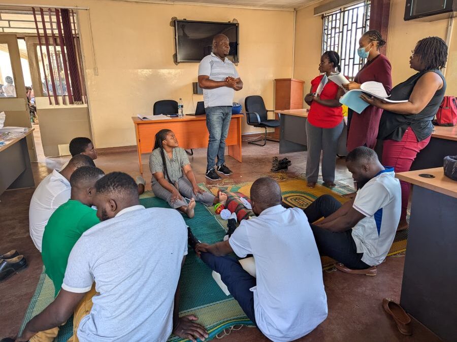 Samuel stands at the front of a room leading a training workshop. Participants are arranged in a loose circle around Sam, some are seated on the floor, while others stand.