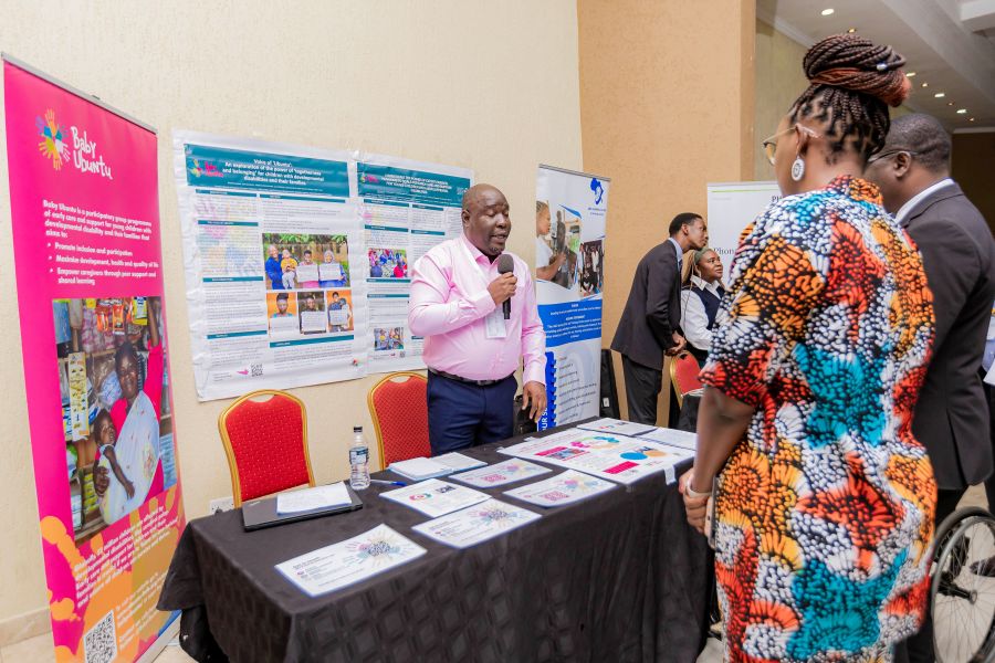 A man holding a microphone stands behind a table. Behind him are posters from Baby Ubuntu in bright colour. The man is talking to two people who stand in front of the table, they look at the man in an engaging way. 